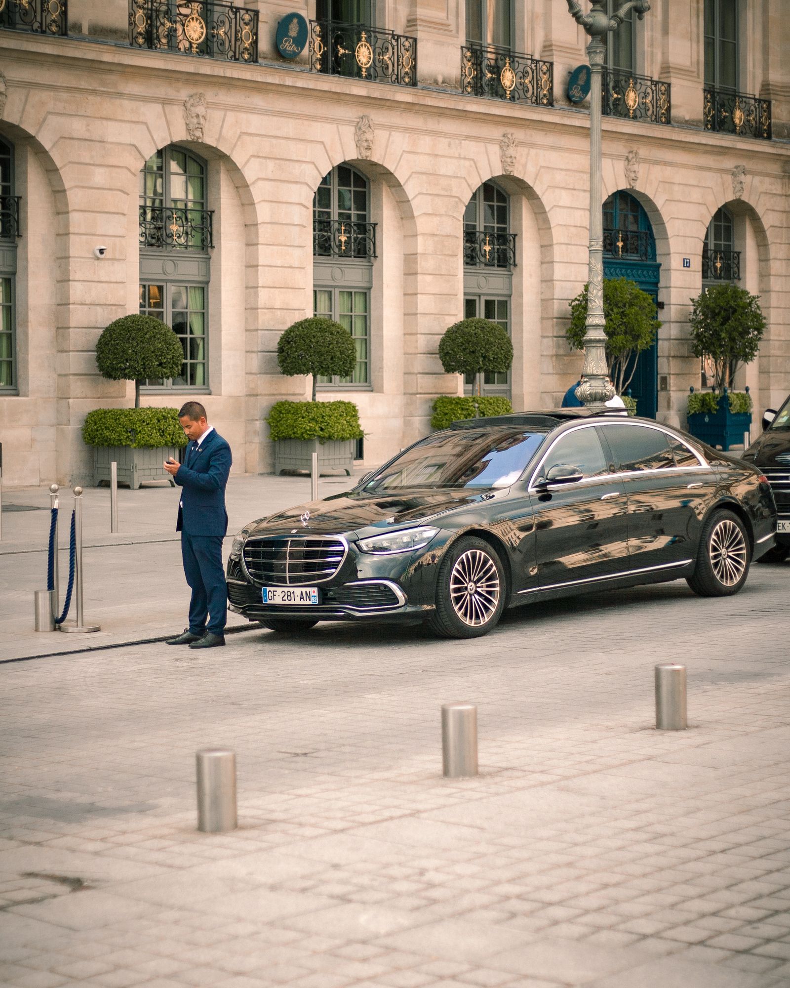A driver in a suit waiting beside a black car outside a hotel